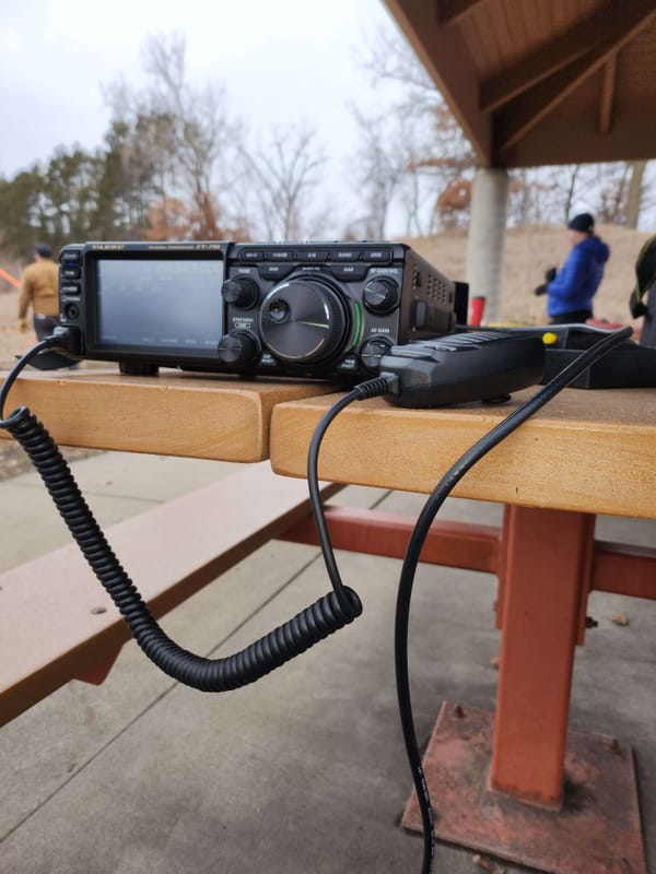 An FT-710 radio and microphone set up on a metal picnic table; Background, other participants and the park itself.